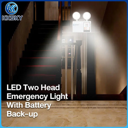 KKSKY LED two-head emergency light with battery backup shown mounted on a staircase landing in a real interior environment, with bright beams of light projecting outward. The product itself is white and mounted above the stairs.